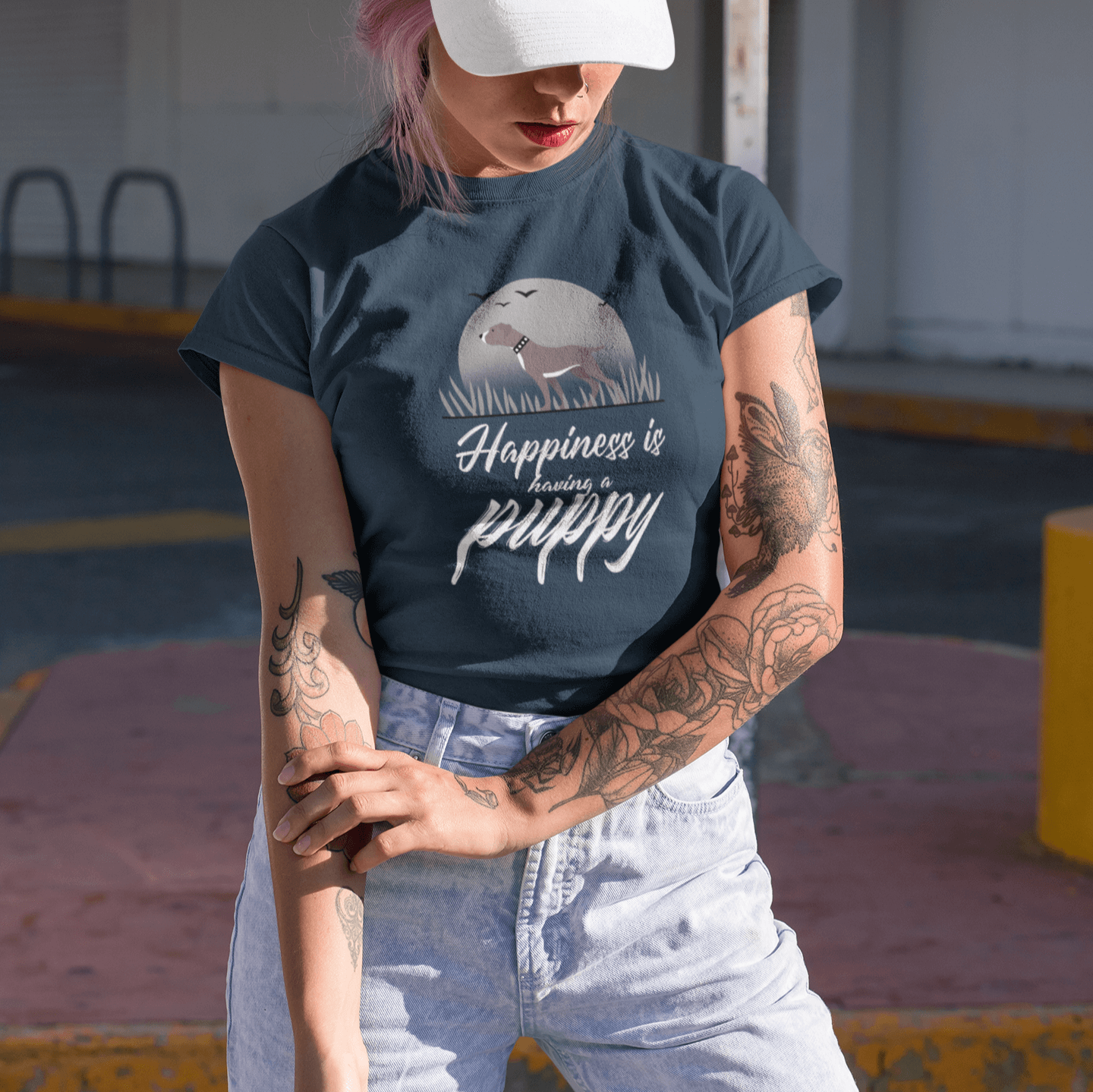 Stylish woman wearing a navy T-shirt with a Pitbull puppy design and white text 'Happiness is having a puppy,' paired with light denim jeans and a white dad hat, posing outdoors under the sunlight