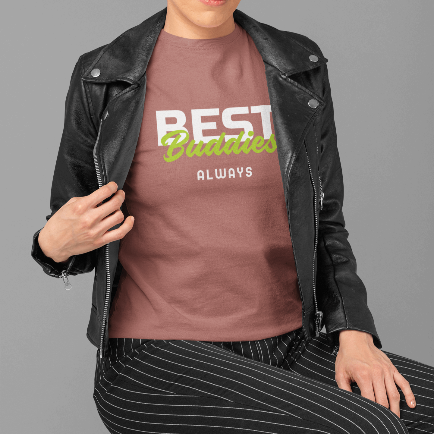 A woman wearing a dusty pink "BEST Buddies ALWAYS" friendship-themed T-shirt, paired with a black leather jacket and striped pants, sitting on a wooden chair with a black headscarf, showcasing a unique and stylish look.