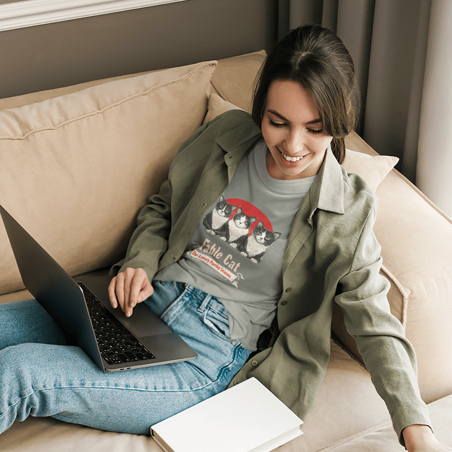 Young woman lounging on a beige sofa, working on a laptop, wearing a gray T-shirt with the 'Fable Cat' design featuring three cats and a red backdrop.