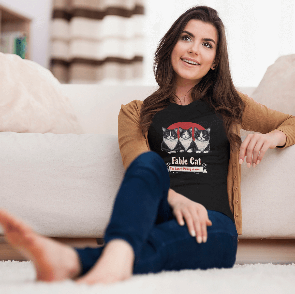 Woman sitting on the floor, smiling, wearing a black T-shirt with 'Fable Cat' design featuring three cats and a red backdrop.