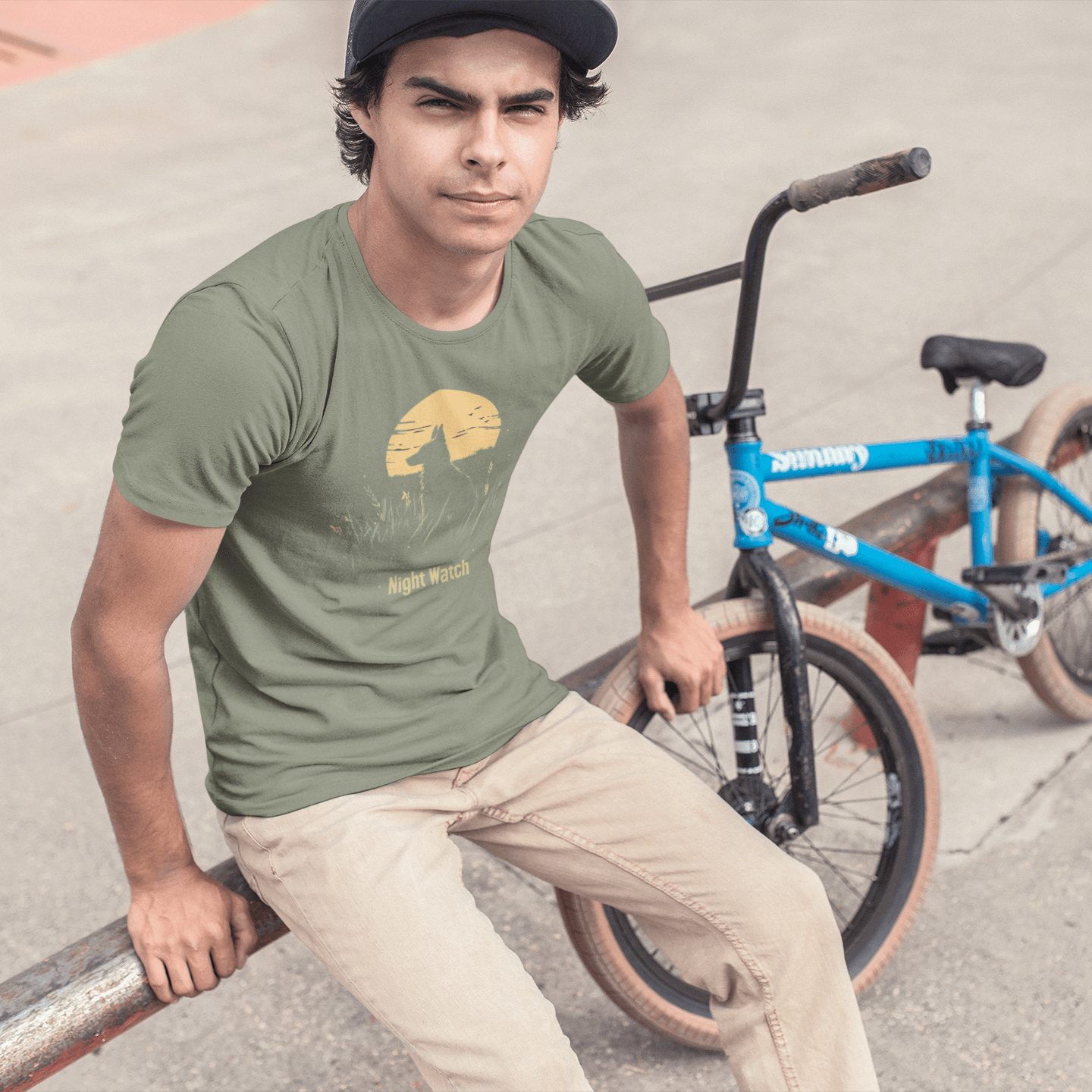 A man wearing a "Night Watch" T-shirt sits on a skatepark railing with a blue bicycle beside him. The design features a dog keeping vigil in tall grass under a full moon, conveying a cool and protective vibe.