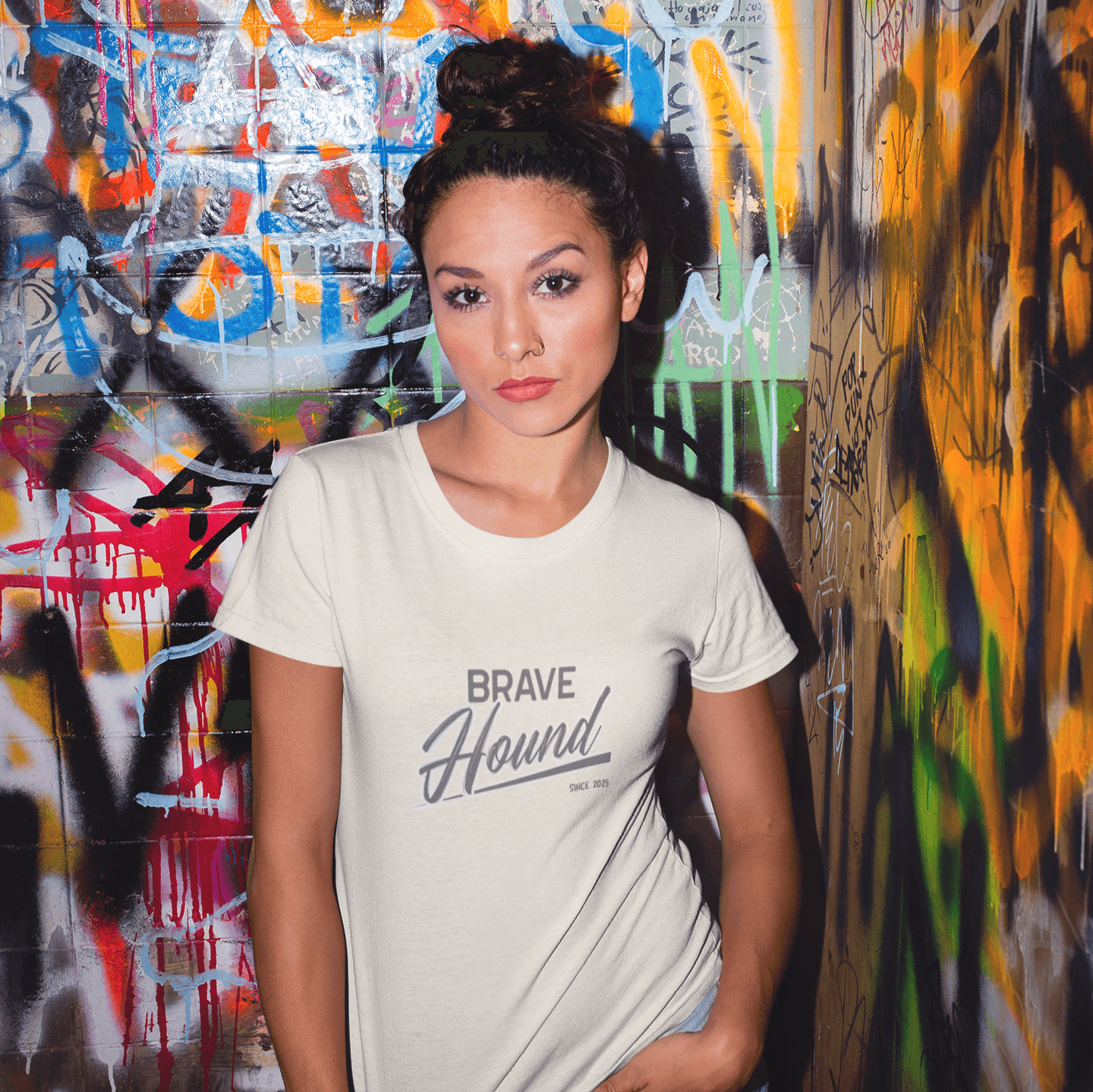 A woman wearing a cream-colored T-shirt with 'Brave Hound' text design, posing confidently in front of a vibrant graffiti-covered wall, styled with light denim shorts.