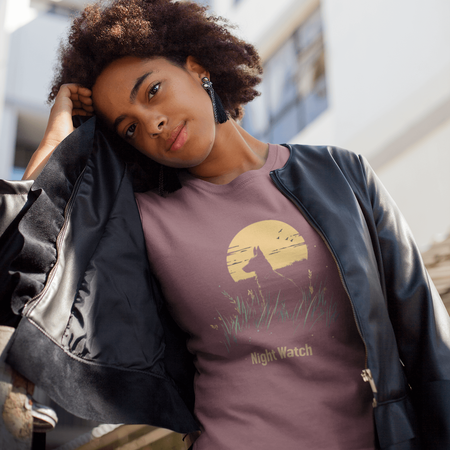 A woman wearing a "Night Watch" T-shirt paired with a black leather jacket stands in front of a modern building. The T-shirt design features a dog keeping vigil in tall grass under a full moon, evoking a cool and resolute vibe.