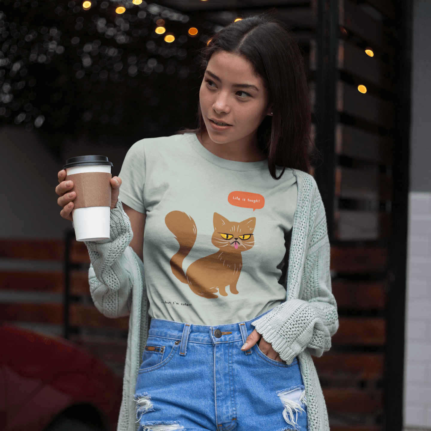 A mockup of a young woman outdoors holding a coffee cup, wearing a light green T-shirt with a design of a grumpy brown cat saying 'Life is tough!' and the caption '...but I'm cuter!' The T-shirt pairs with ripped denim shorts and a mint green cardigan.