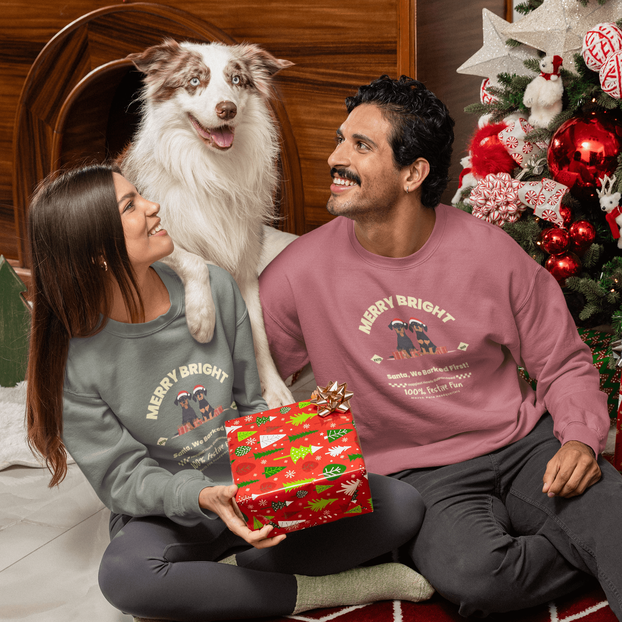 Man and woman in festive sweatshirts with a dog, smiling and holding a Christmas gift by a decorated tree.